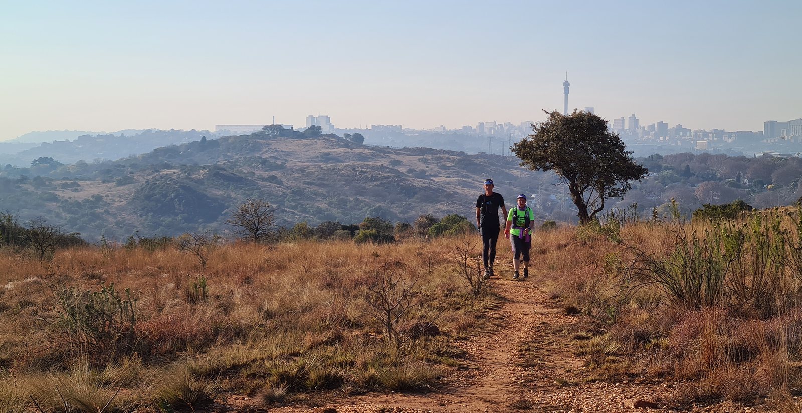 Two runners coming up a rocky trail through winter grass, the Johannesburg skyline on the horizon behind them